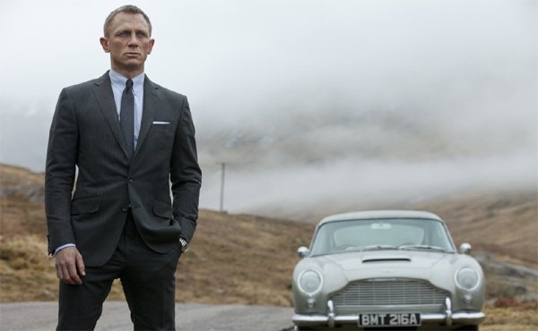 Дэниел Крейг в тёмном костюме рядом с серебристым Aston Martin на фоне туманного пейзажа в фильме «Скайфолл»