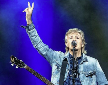 Продюсер четырёх фильмов о Beatles объяснила, как смотреть все истории в кинотеатрах - 1 Пол Маккартни в джинсовой куртке поёт в микрофон и поднимает руку со знаком мира на концерте