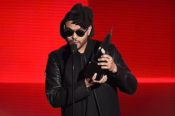The Weeknd в солнцезащитных очках держит награду у микрофона на красном фоне